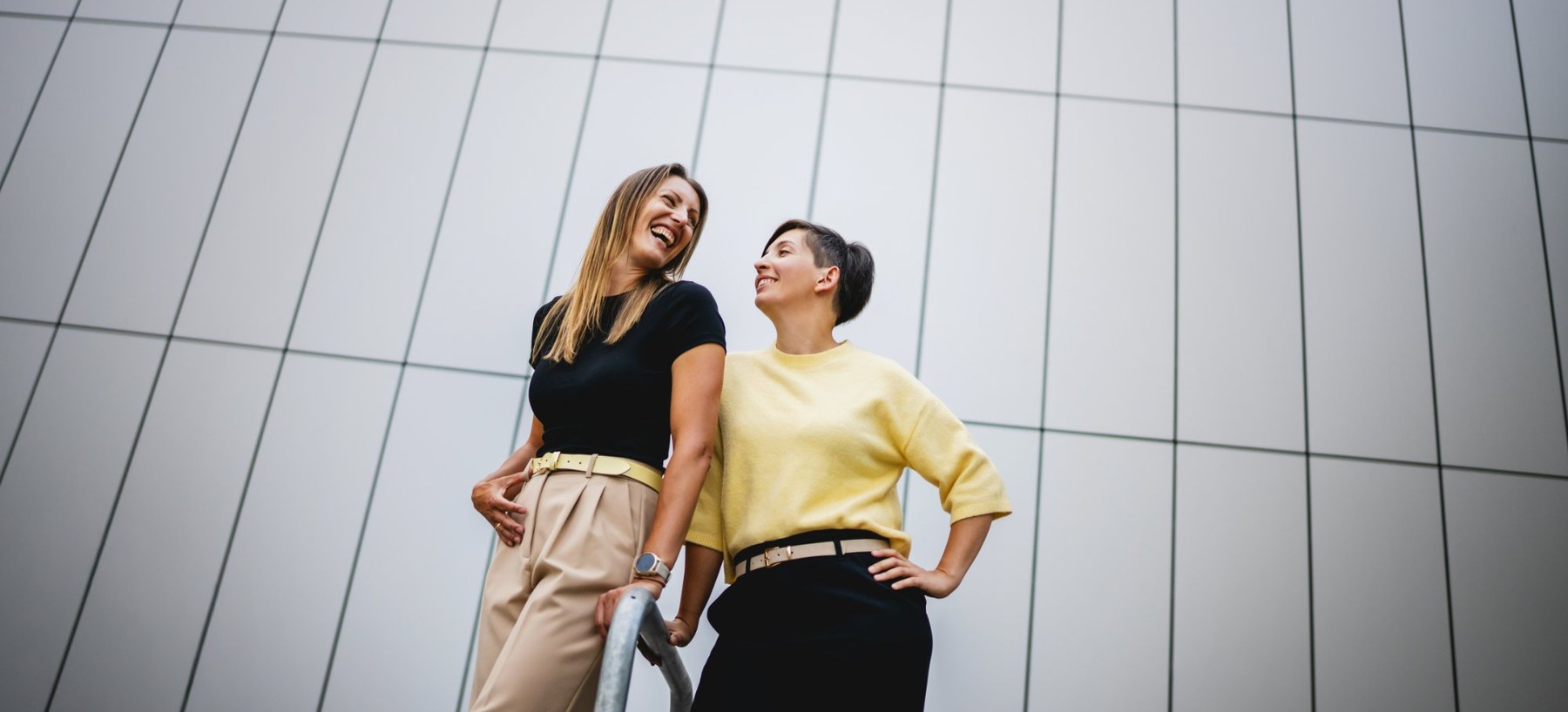 Eva Sturm und Maja Tuder stehen lachend vor einer hellen Gebäudefassade.